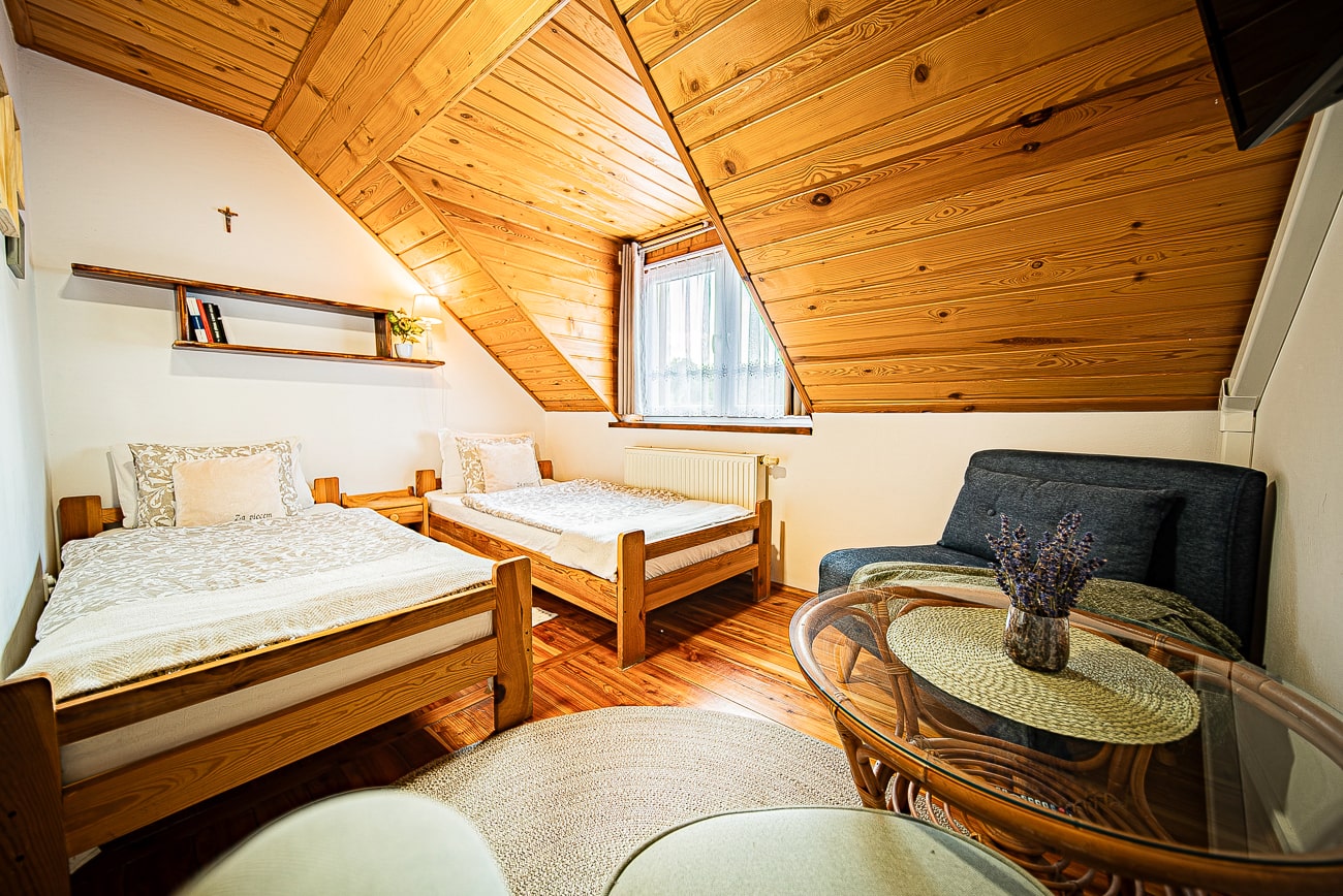 Agroturystyka Za Piecem – Przytulna sypialnia na poddaszu w agroturystyce dla rodziny ze skośnym drewnianym sufitem, dwoma pojedynczymi łóżkami, małą sofą, szklanym stolikiem kawowym z kwiatami i regałem nad łóżkami. Przez okno wpada światło słoneczne.