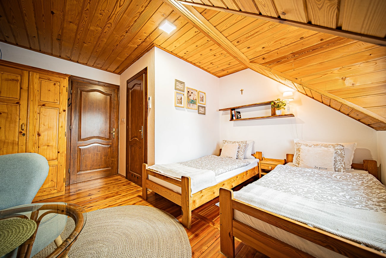 Agroturystyka Za Piecem – Przytulna sypialnia na poddaszu z drewnianymi ścianami i sufitem, idealna na wypoczynek na wsi, wyposażona w dwa pojedyncze łóżka z białą pościelą, drewnianą szafę, półki z dekoracjami i okrągły tkany dywan na drewnianej podłodze.