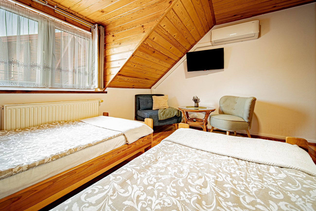 Agroturystyka Za Piecem – Przytulna sypialnia na poddaszu idealna na noclegi dla rodzin z dziećmi, wyposażona w dwa pojedyncze łóżka z wzorzystymi narzutami, skośny drewniany sufit, okno z koronkowymi zasłonami, kaloryfer, mały stolik, wyściełane krzesło, szarą sofę, telewizor na ścianie i klimatyzację.