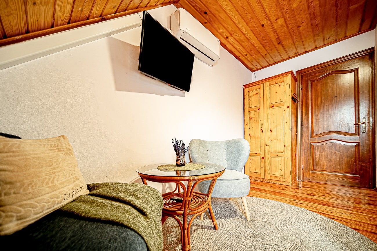 Agroturystyka Za Piecem – Przytulny pokój z drewnianym sufitem i podłogą, szarym fotelem, małym okrągłym stolikiem z wazonem, sofą z poduszką i kocem, drewnianą szafą, telewizorem na ścianie i klimatyzacją - idealny na agroturystykę dla rodziny lub wypoczynek na wsi.