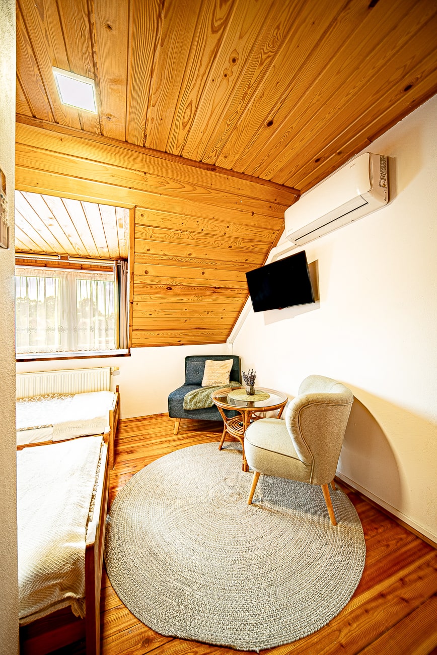 Agroturystyka Za Piecem – Przytulny pokój z drewnianym sufitem i podłogą, idealny na agroturystykę dla rodziny. Znajduje się w nim łóżko, dwa fotele, stolik boczny, telewizor na ścianie, klimatyzator pod skośnym sufitem i światło słoneczne wpadające przez okno.