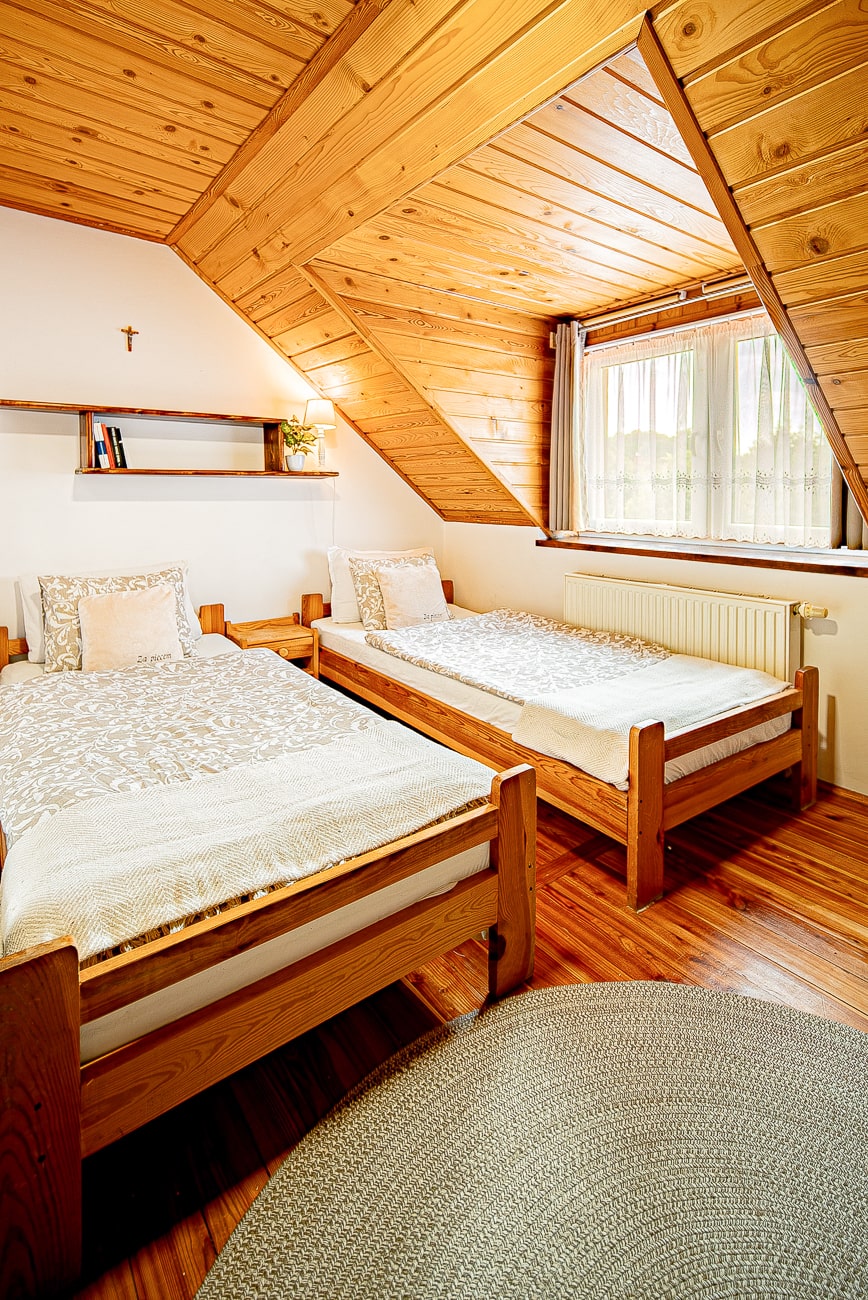 Agroturystyka Za Piecem – Przytulna sypialnia na poddaszu z drewnianymi ścianami i sufitem, dwoma pojedynczymi łóżkami z neutralną pościelą, oknem z przezroczystymi zasłonami, małą półką z rośliną i krzyżem nad łóżkami oraz okrągłym tkanym dywanem - idealnym do agroturystyki dla rodzin poszukujących wypoczynku na wsi.