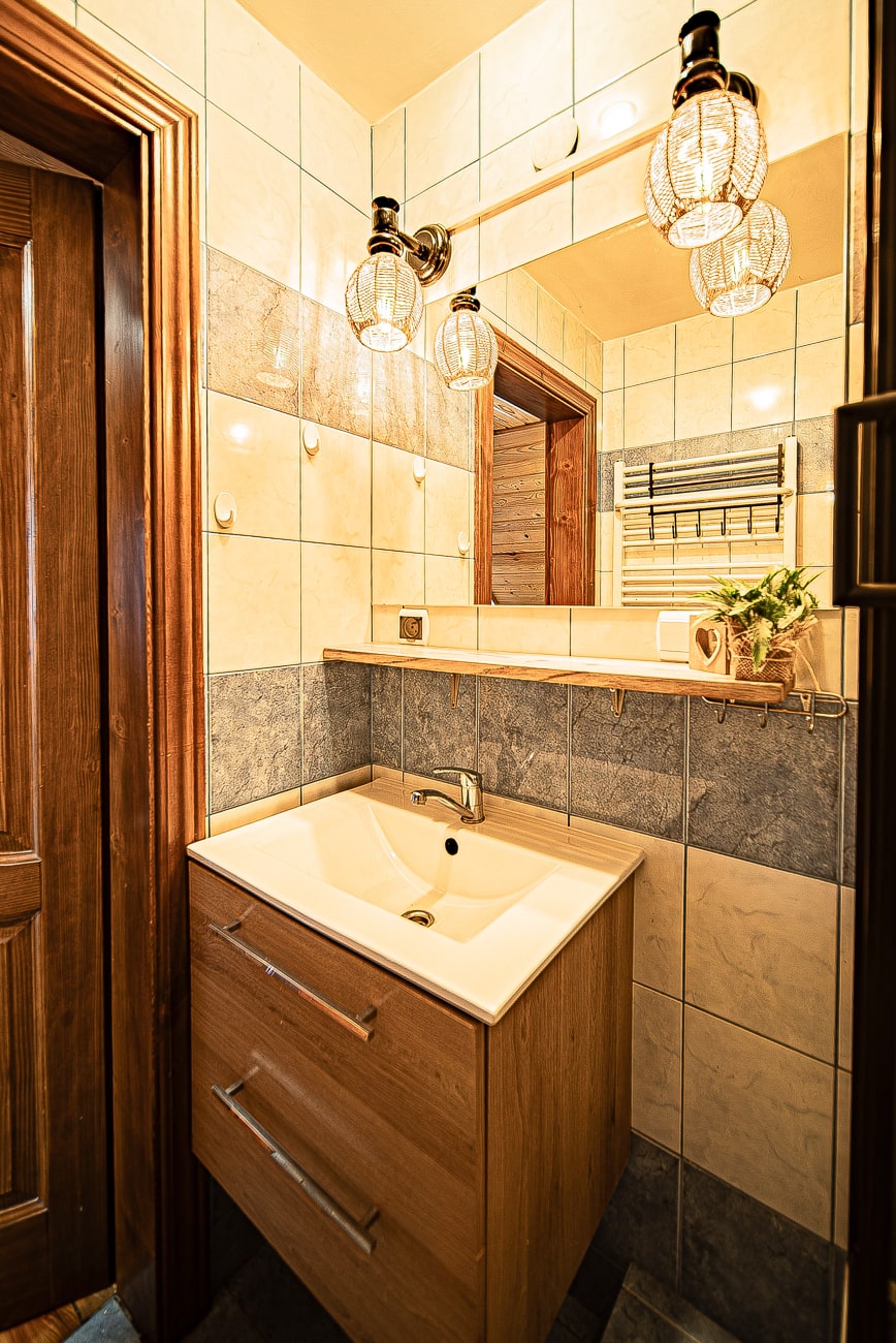 Agroturystyka Za Piecem – Mała, nowoczesna łazienka z wyłożonymi kafelkami ścianami, drewnianą toaletką z umywalką, dużym lustrem, lampkami na ścianie, wieszakiem na ręczniki i rośliną doniczkową na blacie - idealna do spa na wsi lub wypoczynku na wsi.