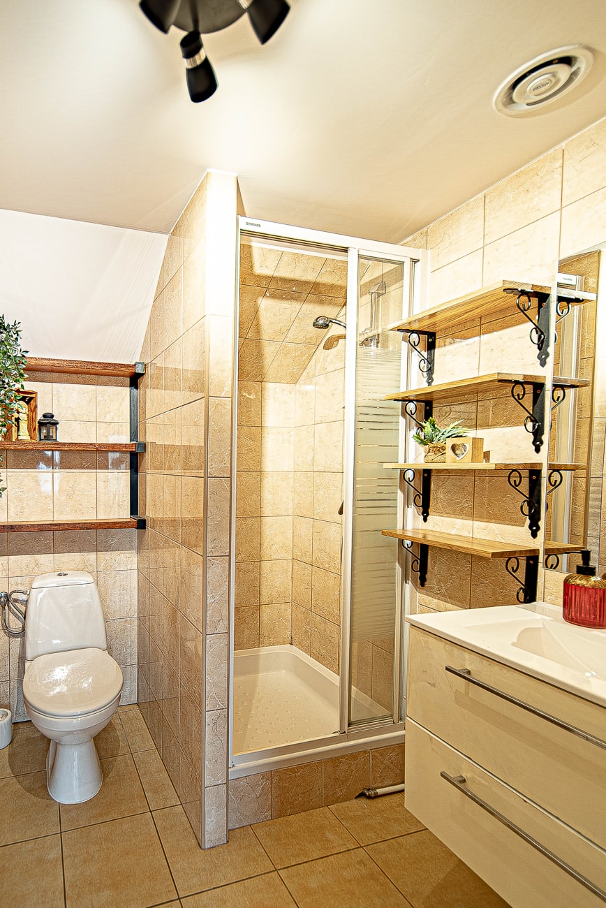Agroturystyka Za Piecem – Nowoczesna łazienka z beżowymi płytkami, szklaną kabiną prysznicową, białą toaletą, wiszącymi półkami z roślinami i dekoracjami oraz białą toaletką z szufladami - idealna do relaksu po dniu spędzonym w spa na wsi lub agroturystyce z dziećmi.