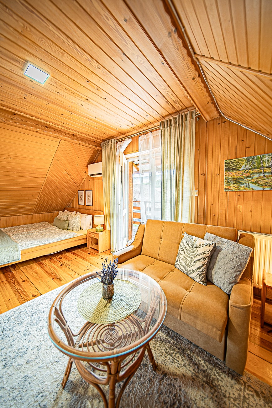 Agroturystyka Za Piecem – Przytulny drewniany pokój z brązową sofą, szklanym stolikiem kawowym i łóżkiem w rogu. Światło słoneczne wpada przez przezroczyste zasłony, a na stole stoi mały wazon z lawendą. Idealny dla agroturystyki dla rodzin poszukujących ciepłej, przytulnej atmosfery z naturalnymi drewnianymi ścianami i podłogą.