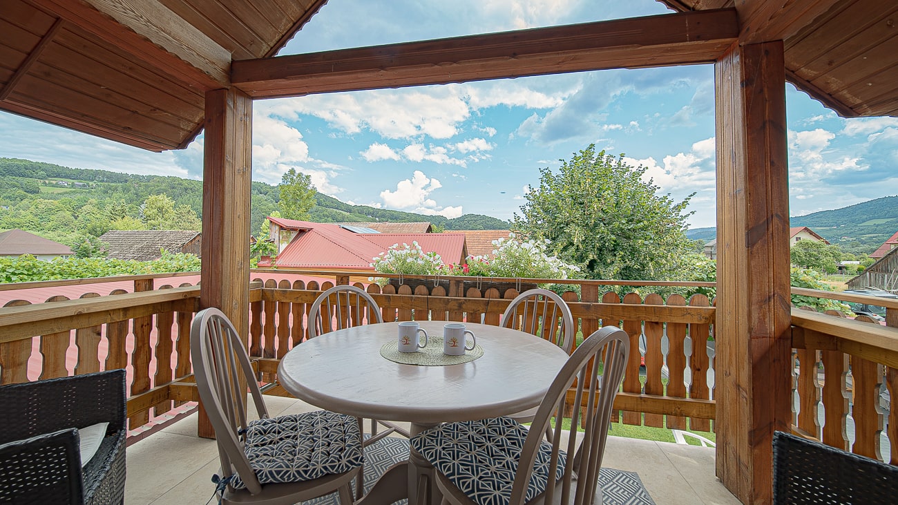 Agroturystyka Za Piecem – Zadaszony drewniany balkon z okrągłym stołem i czterema krzesłami wychodzi na domy z czerwonymi dachami, zielone drzewa i odległe wzgórza pod częściowo zachmurzonym niebem. Idealny na wypoczynek na wsi i noclegi dla rodzin z dziećmi. Na stole stoją trzy kubki.