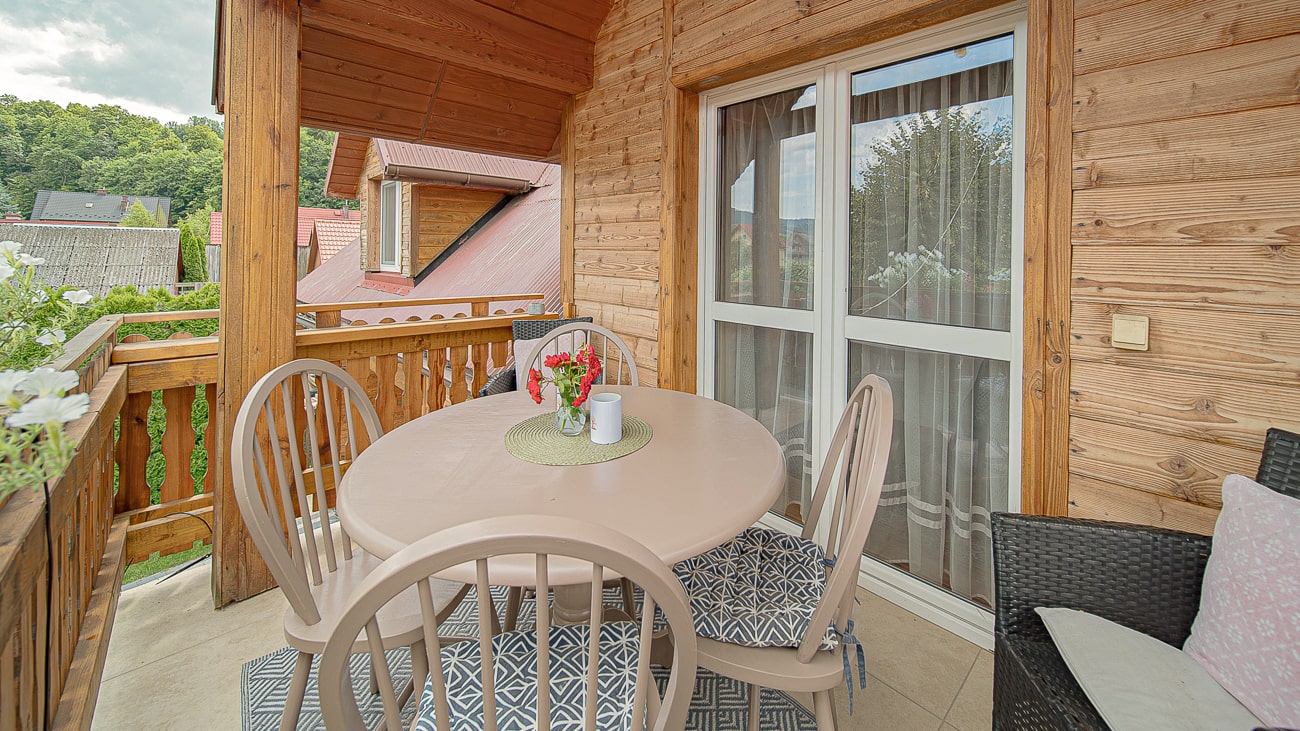Agroturystyka Za Piecem – Przytulny drewniany balkon idealny na agroturystykę z dziećmi, wyposażony w okrągły stół, cztery krzesła, wazon z czerwonymi i białymi kwiatami oraz wyściełaną ławkę. Przesuwane szklane drzwi prowadzą do środka, z zielenią za drewnianą balustradą.
