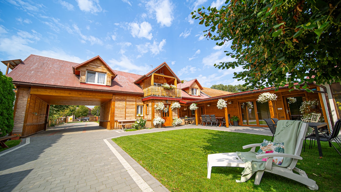 Agroturystyka Za Piecem – Duży drewniany dom z czerwonymi dachami, balkonami i wiszącymi doniczkami stoi obok wybrukowanego podjazdu i zielonego trawnika z leżakami - idealnego do agroturystyki z dziećmi - pod jasnoniebieskim niebem z rozproszonymi chmurami.