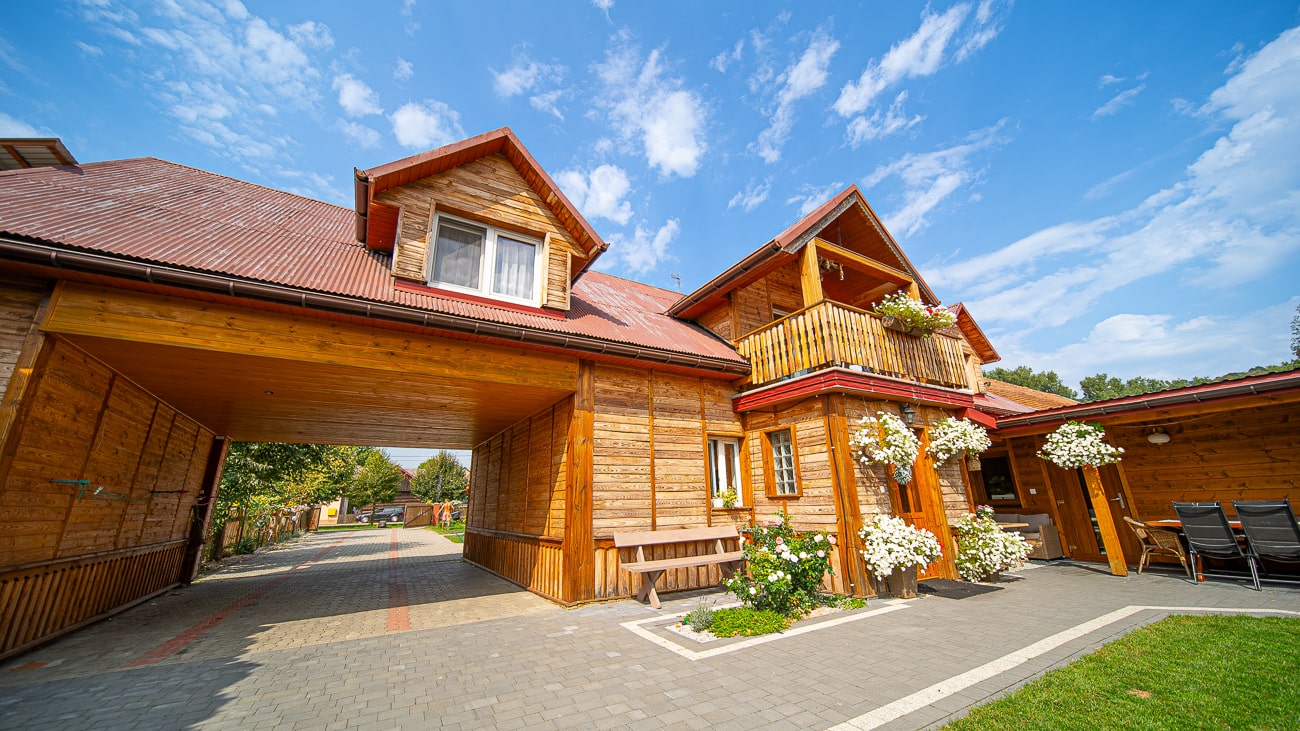 Agroturystyka Za Piecem – Duży drewniany dom z czerwonym dachem, skrzynkami na kwiaty, balkonem i otwartym łukiem nad brukowanym podjazdem oferuje noclegi dla rodzin z dziećmi, ustawione pod jasnym błękitnym niebem z rozproszonymi chmurami.