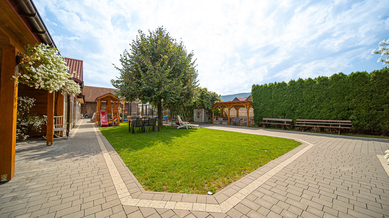 Agroturystyka Za Piecem – Zagospodarowane podwórko z zielonym trawnikiem, wybrukowanym chodnikiem, ławkami i drewnianą altaną - idealne na wypoczynek na wsi. Na podwórku rosną drzewa i krzewy, a pod częściowo zachmurzonym niebem stoją budynki z drewnianym sidingiem.