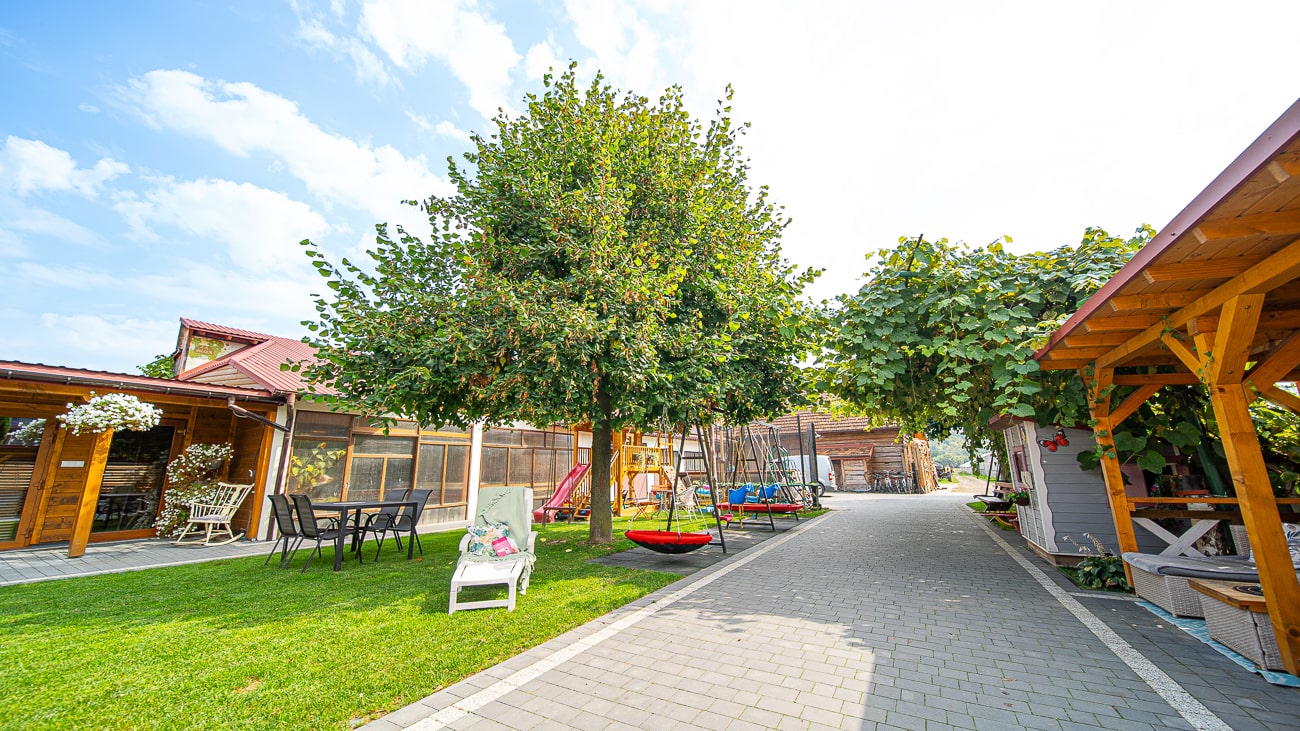 Agroturystyka Za Piecem – Słoneczne podwórko z zieloną trawą, meblami ogrodowymi, drzewem z wiszącymi huśtawkami, wybrukowanym chodnikiem i drewnianymi konstrukcjami altan po obu stronach - idealne na agroturystykę dla rodzin szukających wypoczynku na wsi. Teren wygląda czysto i zachęcająco.