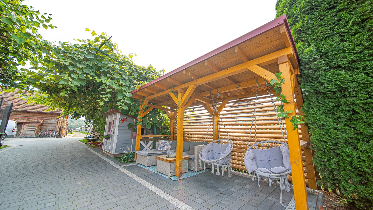 Agroturystyka Za Piecem – Drewniana pergola z czerwonym dachem ocienia dwa wyściełane wiszące krzesła i mały stolik obok brukowanego chodnika - idealne na agroturystykę dla rodziny. W pobliżu znajduje się przytulna szopa i liściaste winorośle, które tworzą przytulną wiejską atmosferę.