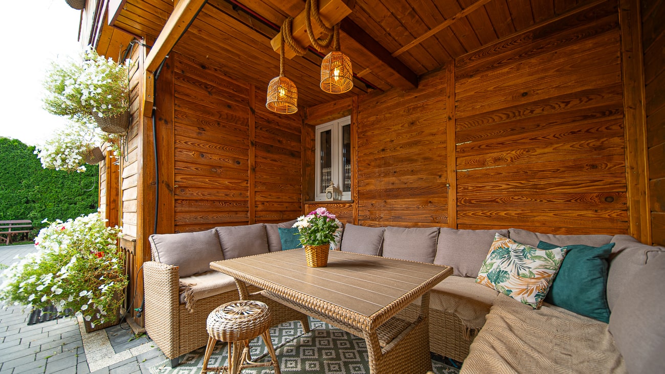 Agroturystyka Za Piecem – Przytulne patio na świeżym powietrzu wyposażone jest w wiklinowy stół i narożną sofę z poduszkami, kwiaty doniczkowe, wiszące lampy i drewniane ściany - idealne do agroturystyki z dziećmi, tworząc ciepłą i zachęcającą rodzinną atmosferę.