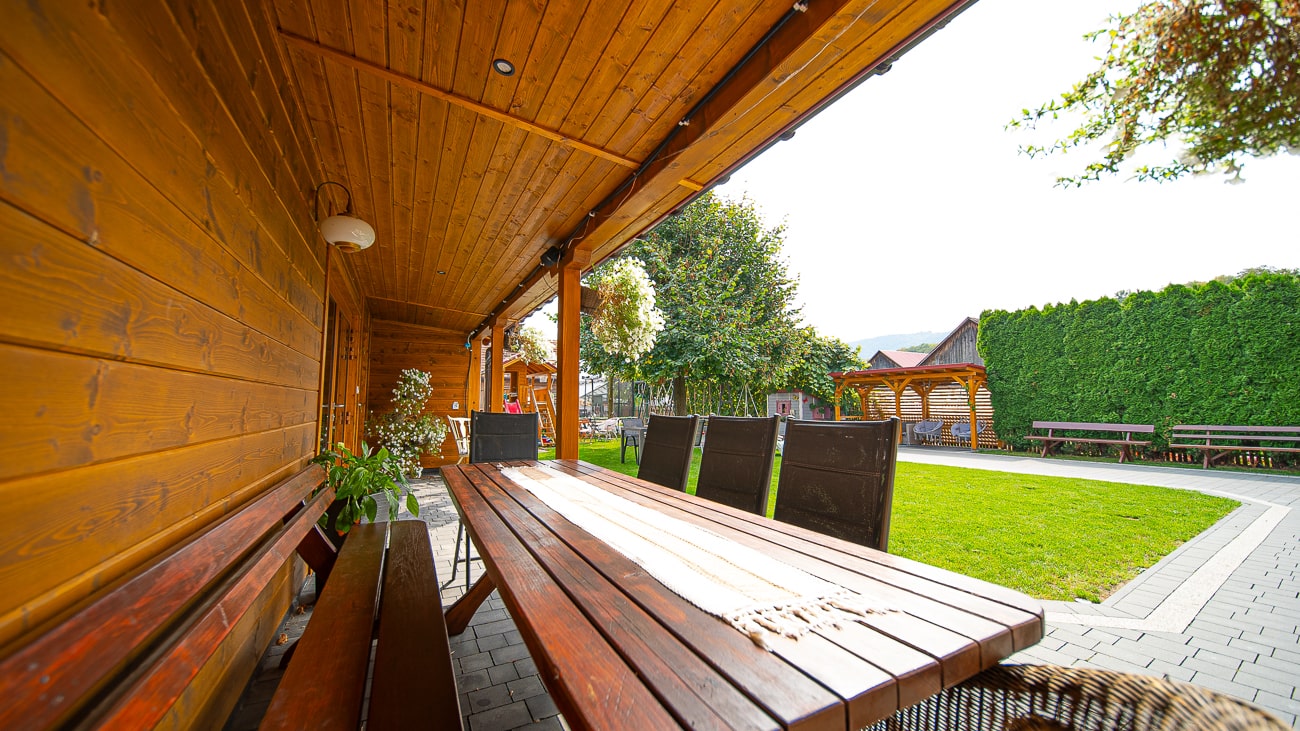 Agroturystyka Za Piecem – Długi drewniany stół z ławkami i krzesłami znajduje się pod zadaszonym patio obok trawnika z drzewami, żywopłotami i ławkami - idealny do agroturystyki dla rodziny w słonecznym przydomowym ogrodzie.