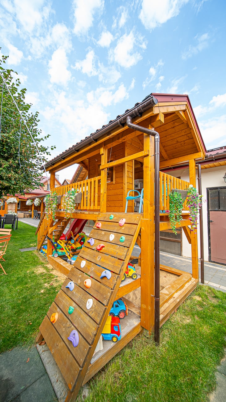 Agroturystyka Za Piecem – Drewniany domek do zabawy z małą werandą, roślinami doniczkowymi, ścianką wspinaczkową i kolorowymi zabawkami pod spodem stoi na zielonej trawie pod czystym, błękitnym niebem - idealny na agroturystykę z dziećmi lub noclegi dla rodzin z dziećmi.