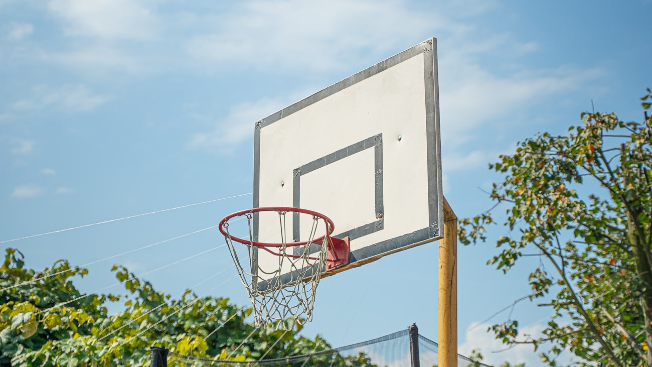 Agroturystyka Za Piecem – Zbliżenie na obręcz do koszykówki na świeżym powietrzu z czerwoną obręczą i linami, na tle zielonych drzew i błękitnego nieba - idealne na aktywny wypoczynek na wsi z rodziną.