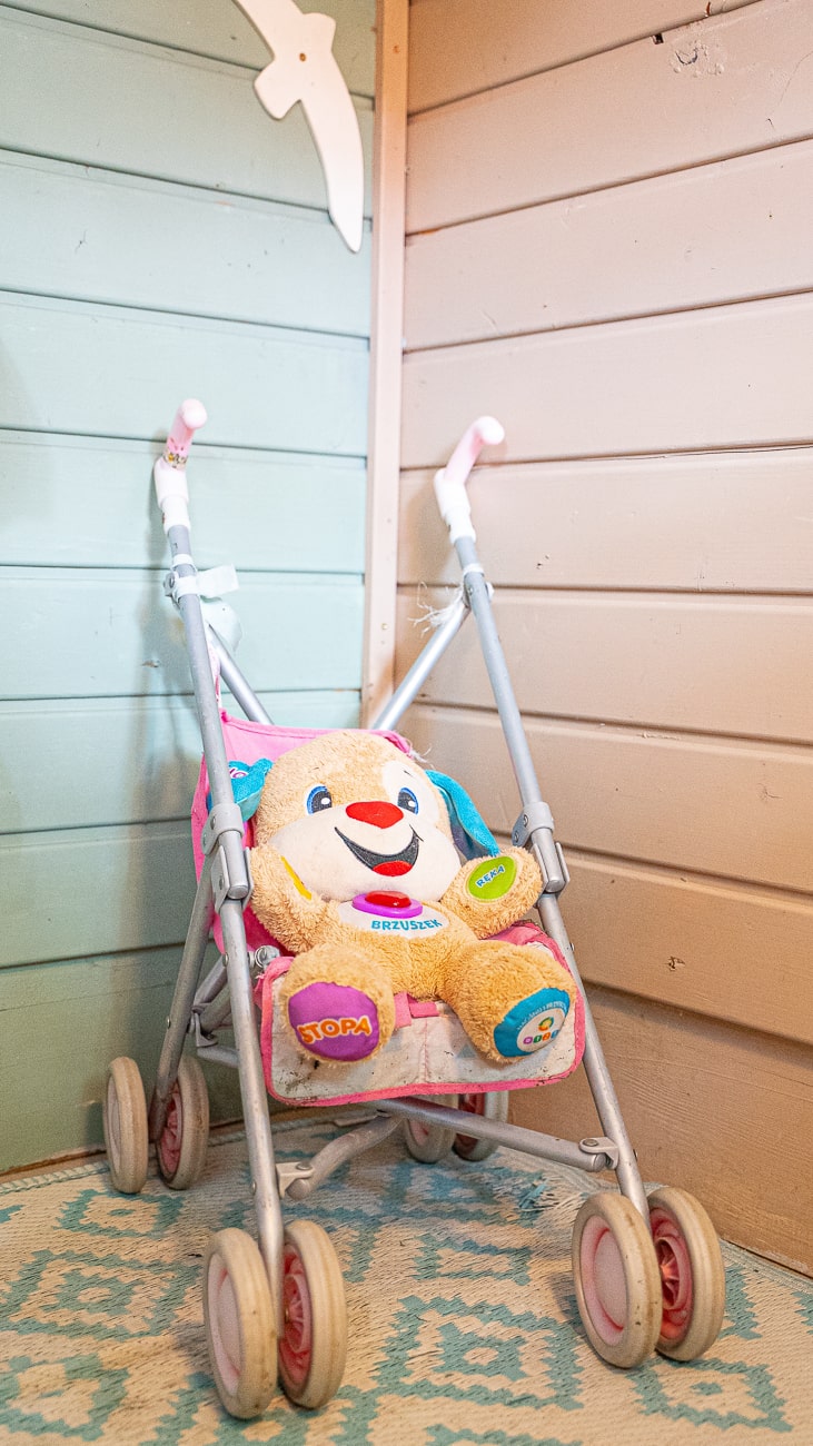 Agroturystyka Za Piecem – Pluszowy miś siedzi w małym różowo-szarym wózku, umieszczonym na niebiesko-białym wzorzystym dywanie obok pastelowych drewnianych ścian - idealny dla rodzin odkrywających agroturystykę ze zwierzętami lub szukających noclegi dla rodzin z dziećmi.