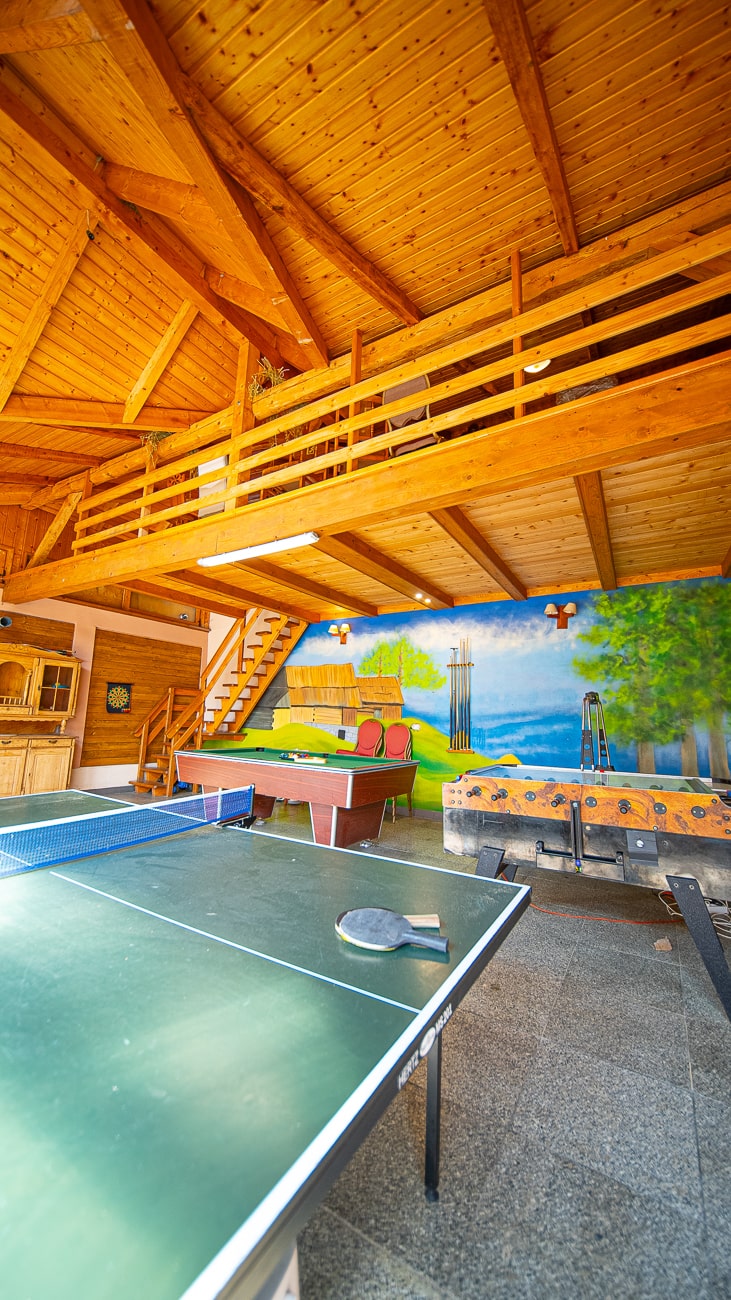 Agroturystyka Za Piecem – Przestronny drewniany pokój gier dla agroturystyki dla rodzin, wyposażony w stół do ping ponga, stół do piłkarzyków, fototapetę z ławkami i drzewami na tylnej ścianie oraz poddasze z drewnianymi balustradami nad nim - idealne na wypoczynek na wsi.