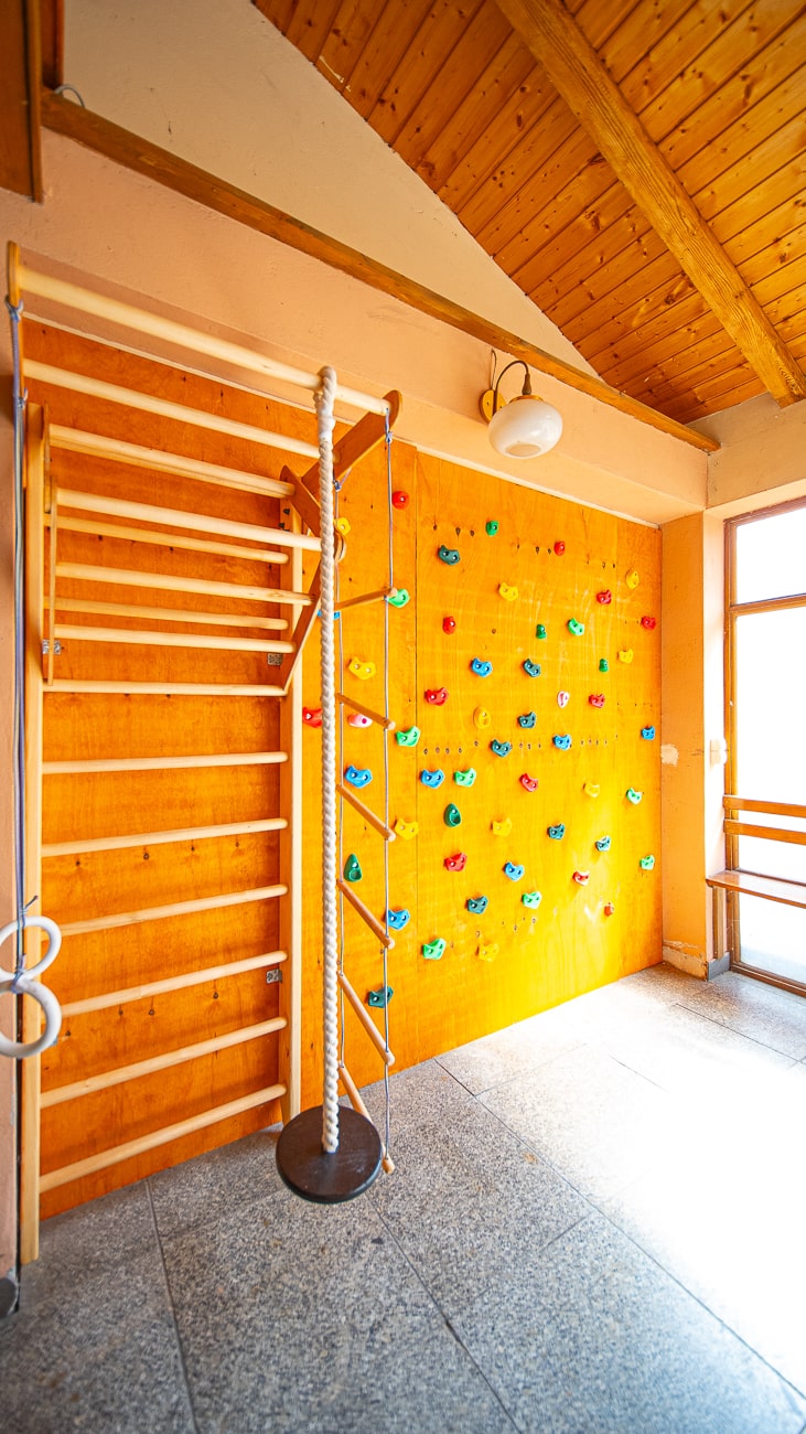 Agroturystyka Za Piecem – Jasna sala gimnastyczna idealna na noclegi dla rodzin z dziećmi, wyposażona w drewnianą ściankę wspinaczkową z kolorowymi uchwytami, linę do wspinaczki, kółka gimnastyczne i drążki ścienne, a wszystko to ustawione na pomarańczowej ścianie pod drewnianym sufitem.