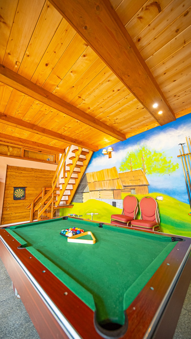 Agroturystyka Za Piecem – Stół bilardowy z bilami ułożonymi do gry znajduje się w przytulnym drewnianym pokoju. Dwa czerwone krzesła, fototapeta ze stodołą i drzewami, tarcza do rzutek i schody tworzą scenerię idealną na agroturystykę z dziećmi lub noclegi dla rodzin z dziećmi.