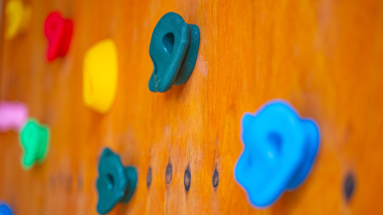 Agroturystyka Za Piecem – Zbliżenie na drewnianą ściankę wspinaczkową z kolorowymi uchwytami, idealną dla rodzin korzystających z noclegów dla rodzin z dziećmi. Uchwyty w kolorach niebieskim, zielonym, żółtym, czerwonym i różowym są rozmieszczone w różnych pozycjach.