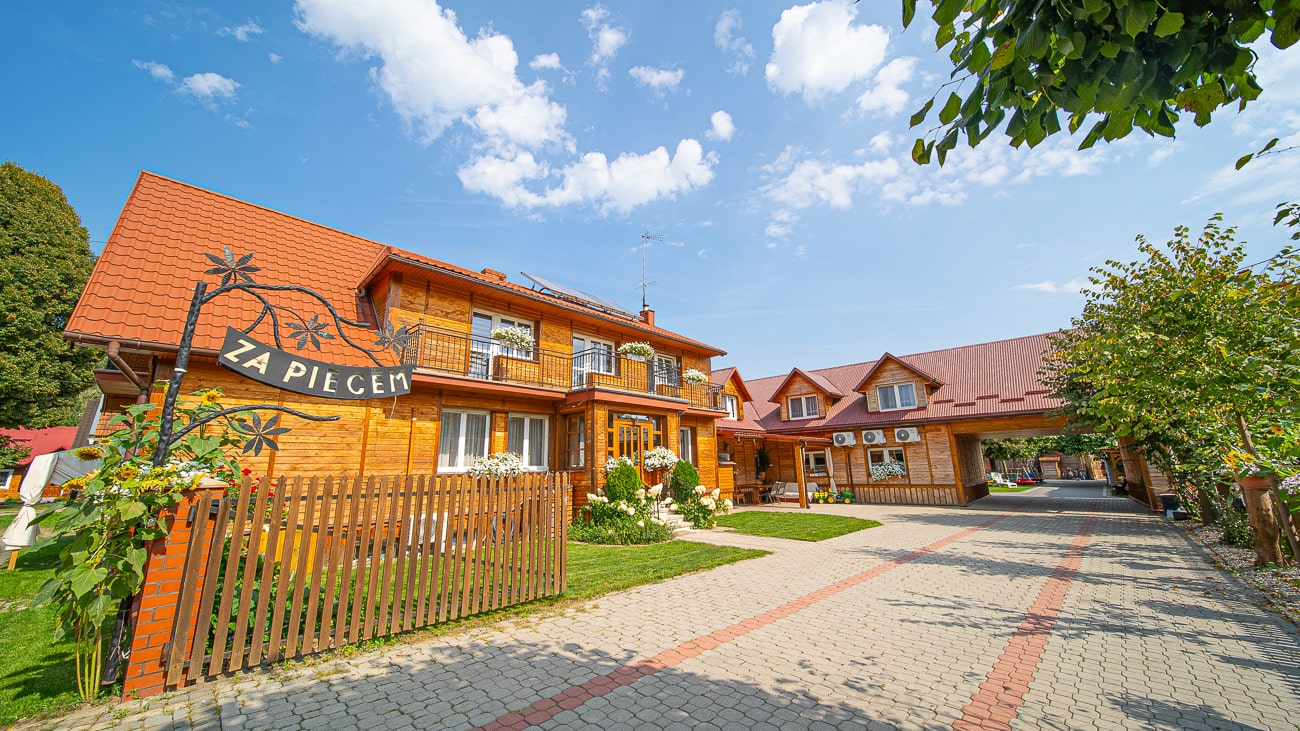 Agroturystyka Za Piecem – Jasny, drewniany pensjonat z czerwonymi dachami i ozdobną bramą Za Piecem stoi obok brukowanego podjazdu, otoczony zielonymi trawnikami i kwitnącymi kwiatami - idealny na agroturystykę z dziećmi pod błękitnym niebem z rozproszonymi chmurami.