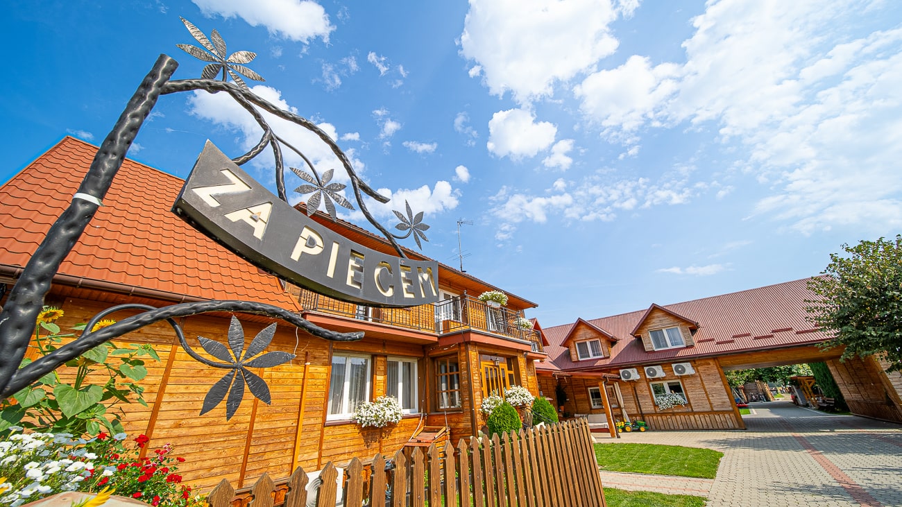 Agroturystyka Za Piecem – Drewniany budynek z czerwonymi dachami i balkonami, otoczony płotem i kwiatami, oferuje agroturystykę ze zwierzętami. Ozdobny metalowy szyld z napisem "ZA PIECE" i motywami kwiatowymi stoi na pierwszym planie pod jasnym, błękitnym niebem.
