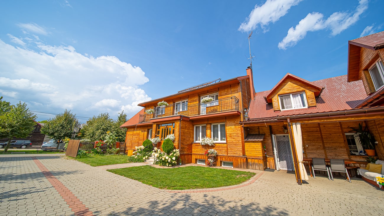 Agroturystyka Za Piecem – Duży drewniany dom z czerwonym dachem stoi pod błękitnym niebem z białymi chmurami. Otoczona zieloną trawą i drzewami agroturystyka dla rodzin oferuje balkony z kwiatami i wybrukowany podjazd, idealny na relaksujące spa na wsi.