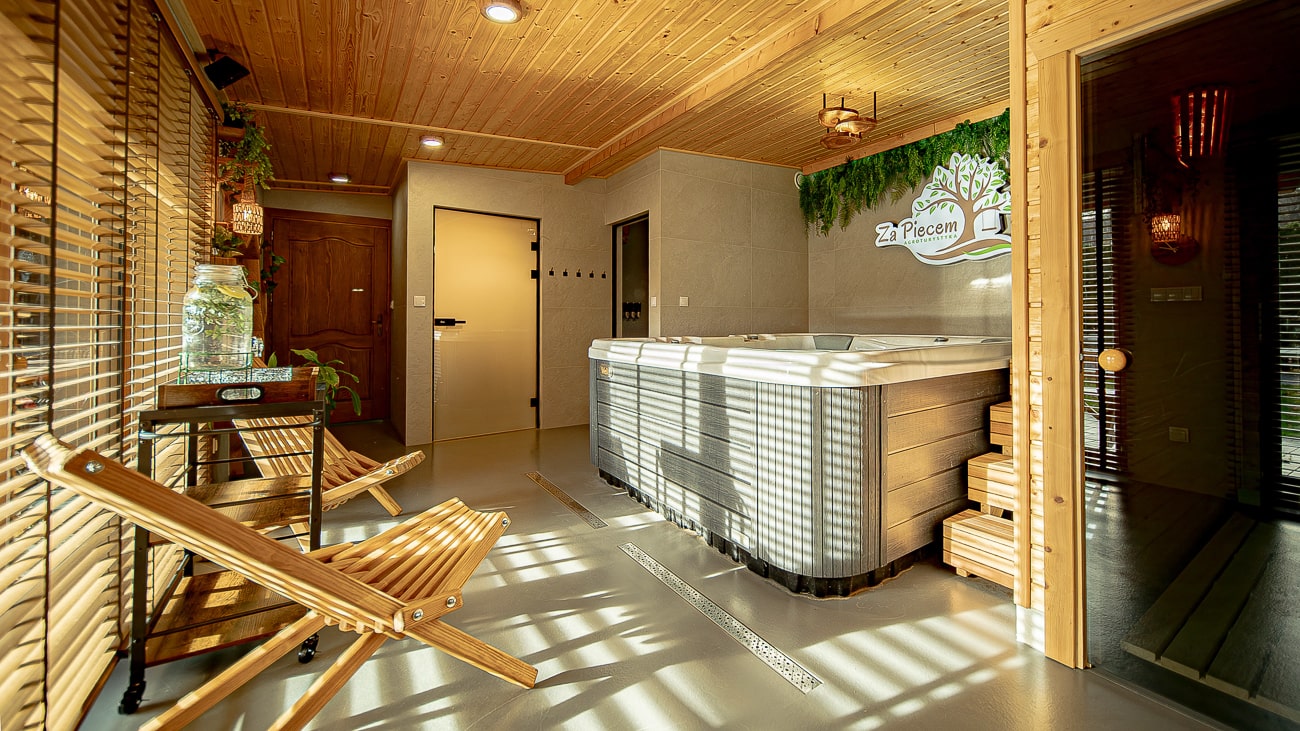 Agroturystyka Za Piecem – Przytulne wewnętrzne spa na wsi z drewnianym sufitem i ścianami, wanną z hydromasażem, dwoma drewnianymi leżakami, roślinami doniczkowymi i światłem słonecznym wpadającym przez żaluzje okienne, tworząc pasiaste cienie na podłodze i meblach.