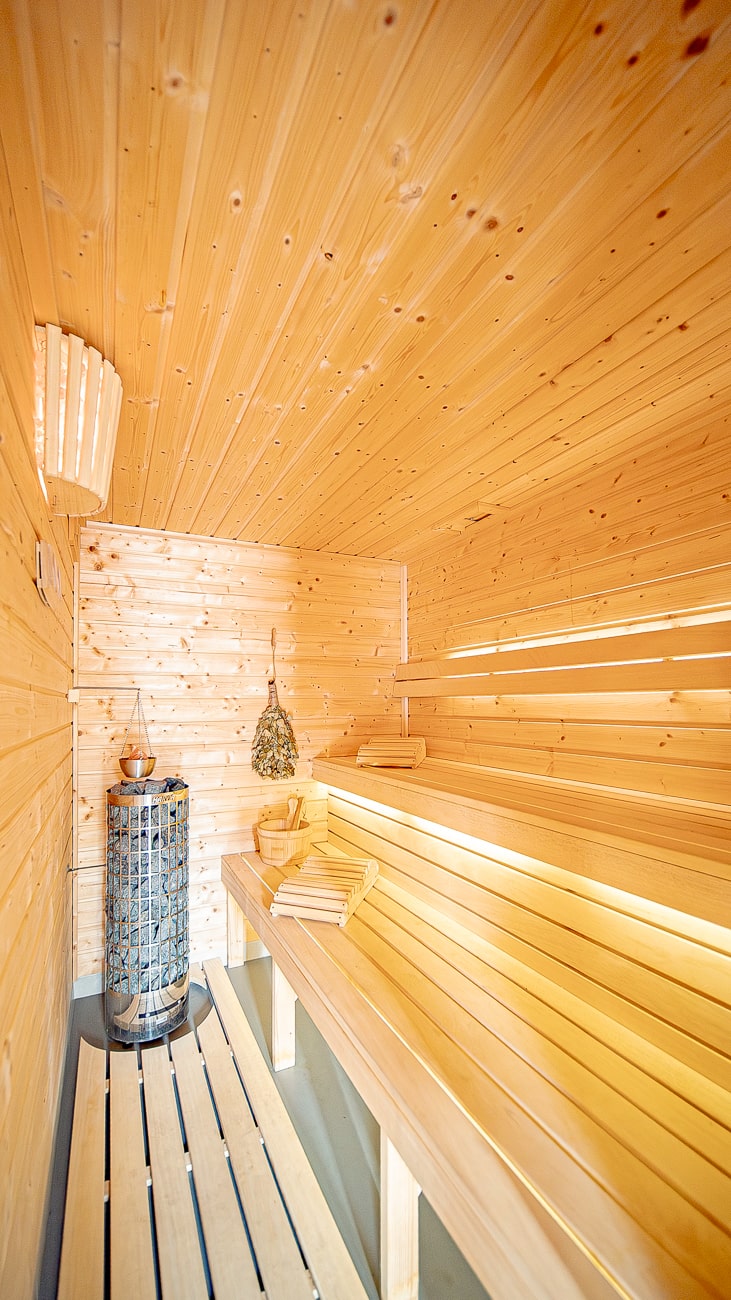 Agroturystyka Za Piecem – Jasne, drewniane wnętrze sauny z piętrowymi ławami, metalowym piecem, wiadrem z chochlą i pękami suszonych liści wiszącymi na ścianie. Ciepłe oświetlenie podkreśla przytulną atmosferę - idealną na agroturystykę z dziećmi i wypoczynek na wsi.