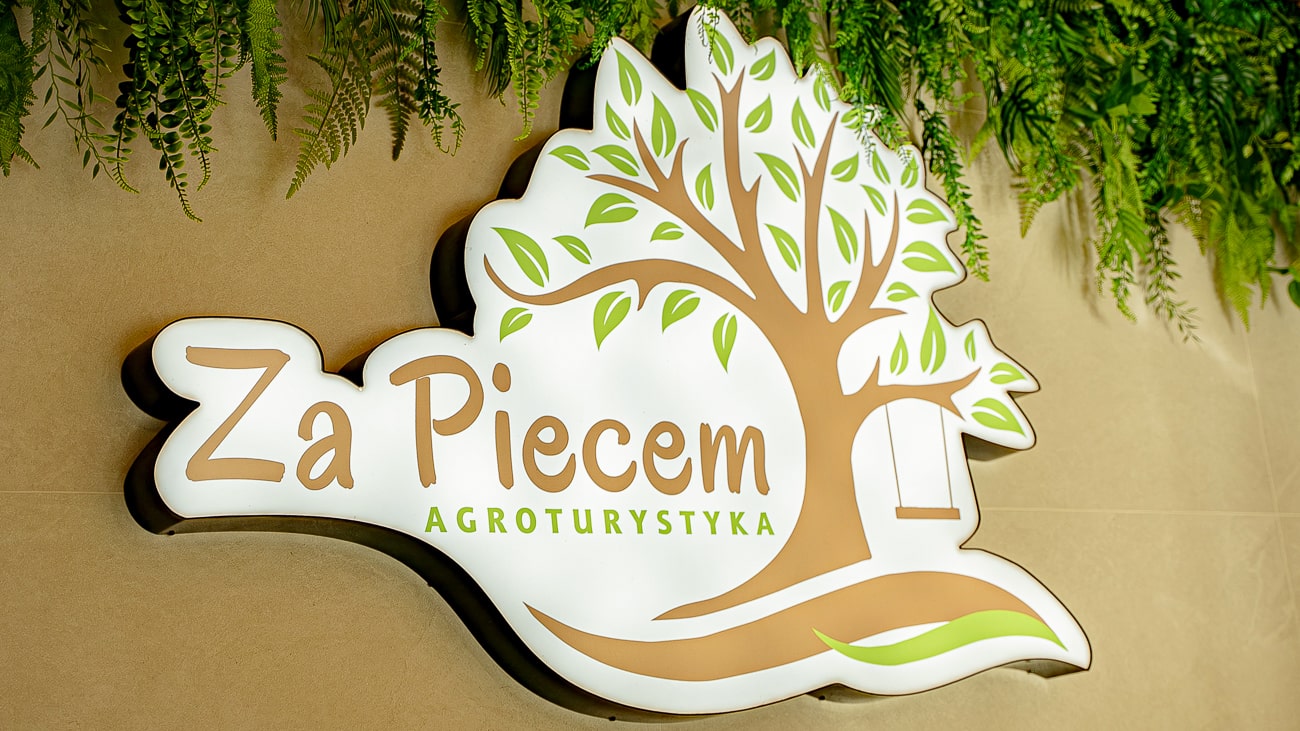 Agroturystyka Za Piecem – Znak z zielono-brązową grafiką drzewa i napisem Za Piecem Agroturystyka na wsi na beżowej ścianie, z zielonymi liściastymi roślinami wiszącymi powyżej - idealny dla rodzin i agroturystyka z dziećmi.