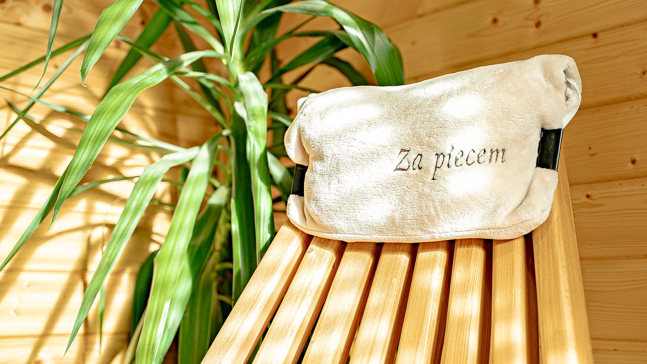 Agroturystyka Za Piecem – Biały ręcznik z wyhaftowanym napisem "Za piecem" spoczywa na drewnianej ławce obok zielonej rośliny liściastej w przytulnym, nasłonecznionym pokoju - idealnym na wypoczynek na wsi lub relaksujące spa na wsi.