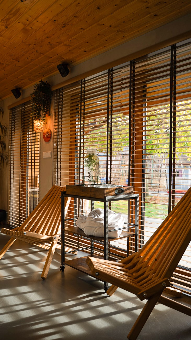 Agroturystyka Za Piecem – Dwa drewniane fotele siedzą przy dużych oknach z poziomymi żaluzjami, rzucając cienie w paski. Pomiędzy krzesłami znajduje się mały stolik z rośliną i ręcznikami. Światło słoneczne wpada do środka, tworząc przytulne miejsce na wypoczynek na wsi lub agroturystykę dla rodziny.