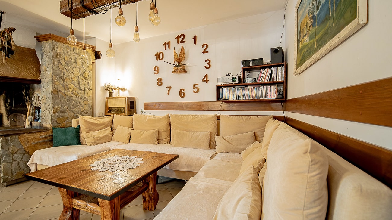 Agroturystyka Za Piecem – Przytulny salon w agroturystyce z dziećmi z dużą beżową rozkładaną sofą, drewnianym stolikiem kawowym, dekoracyjnym zegarem ściennym, kamiennym kominkiem, półkami z książkami i płytami CD oraz obrazem. Ciepłe oświetlenie tworzy przyjazną atmosferę.