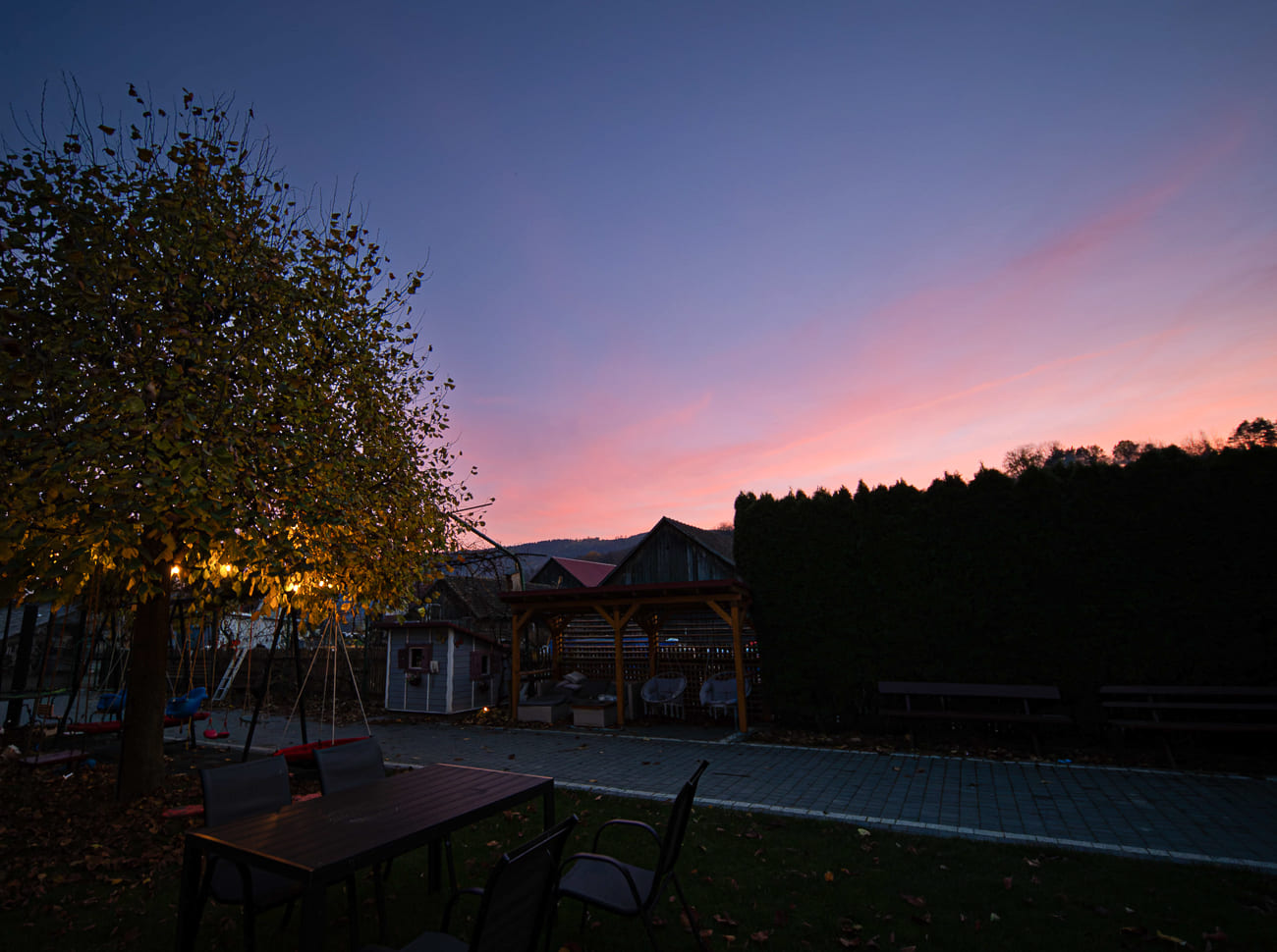 Agroturystyka Za Piecem – Patio o zachodzie słońca ze stołami i krzesłami na trawie, oświetlonym drzewem po lewej stronie, drewnianą altaną pośrodku i ścieżką z ławkami - wszystko idealne na agroturystykę dla rodziny pod niebem w odcieniach różu i błękitu.