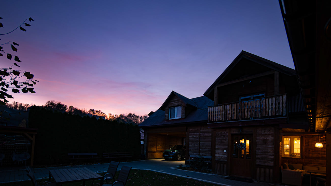 Agroturystyka Za Piecem – Drewniany dom o zmierzchu z ciepłymi światłami świecącymi wewnątrz, idealny na noclegi dla rodzin z dziećmi. Samochód jest zaparkowany pod wiatą, a na pierwszym planie znajdują się meble ogrodowe. Fioletowe i różowe niebo o zachodzie słońca, z drzewami i żywopłotami otaczającymi scenę.