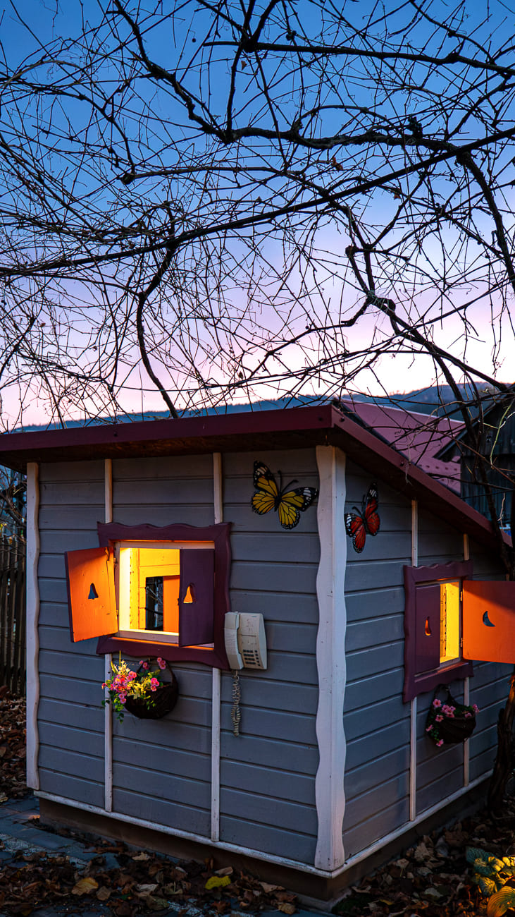 Agroturystyka Za Piecem – Mały, szary drewniany domek z żółtym światłem świecącym z okien stoi pod gołymi gałęziami drzew o zmierzchu. Idealny do agroturystyki z dziećmi, kolorowe dekoracje motyli i skrzynki na kwiaty zdobią jego zewnętrzną część.