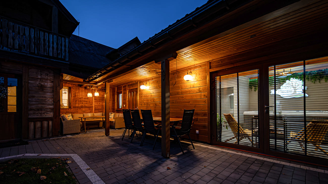 Agroturystyka Za Piecem – Zewnętrzna część wypoczynkowa drewnianego domu o zmierzchu, ciepło oświetlona kinkietami, ze stołem i krzesłami na wybrukowanym patio; przytulne wnętrze widoczne przez przeszklone drzwi. Idealny na agroturystykę z dziećmi lub relaksujące spa na wsi.