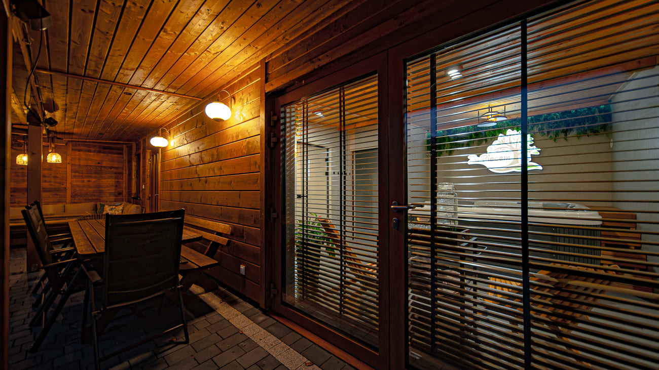 Agroturystyka Za Piecem – Przytulne, zadaszone drewniane patio z ciepłym oświetleniem, stołem i krzesłami oraz szklanymi drzwiami z żaluzjami. Podświetlany szyld prześwituje przez drzwi, oddając zapraszającą atmosferę agroturystyki ze zwierzętami.