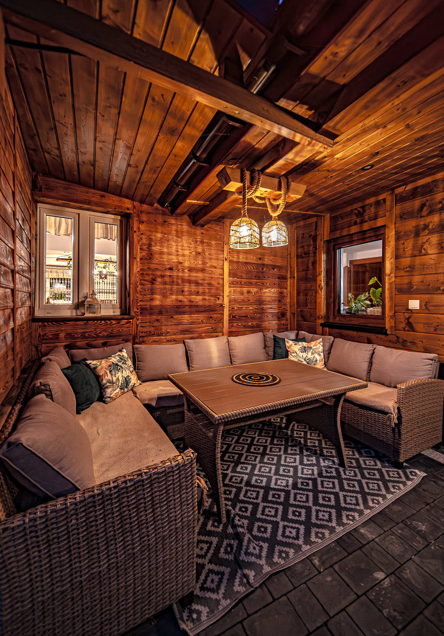 Agroturystyka Za Piecem – Przytulny drewniany domek idealny na wypoczynek na wsi, wyposażony w wiklinową sofę segmentową z wzorzystymi poduszkami, kwadratowy stół i geometryczny dywan. Z sufitu zwisają ciepłe światła, a dwa okna otaczają spokojne widoki na zewnątrz.