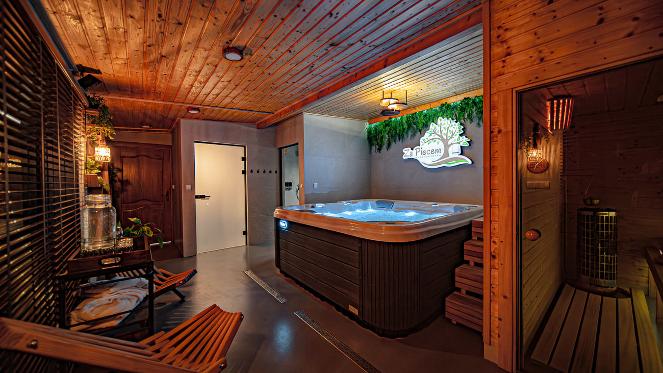 Agroturystyka Za Piecem – Przytulny wewnętrzny pokój spa z drewnianymi ścianami i sufitem wyposażony jest w dużą wannę z hydromasażem, saunę, część wypoczynkową, rośliny i świecący napis "SPA Place". Ciepłe oświetlenie i rustykalne akcenty przywołują urok spa na wsi-idealne noclegi dla rodzin z dziećmi.