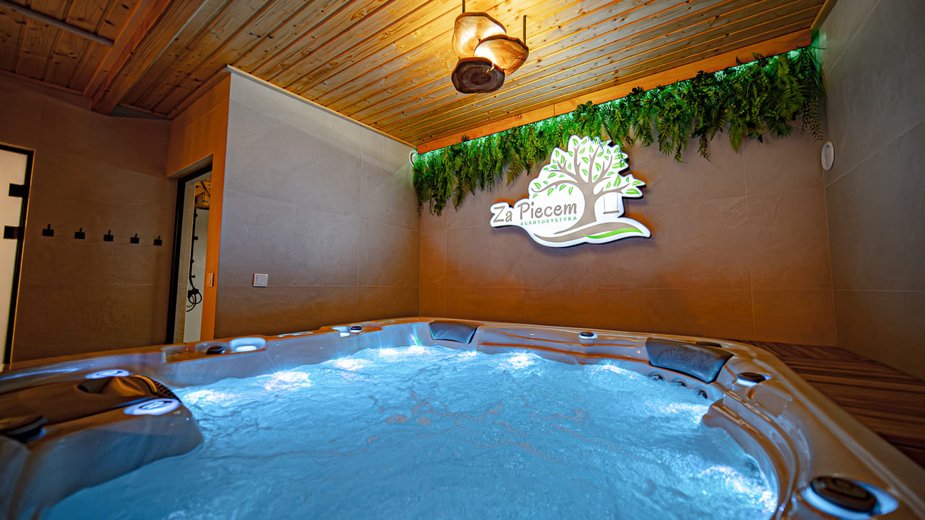 Agroturystyka Za Piecem – Nowoczesna wewnętrzna wanna z hydromasażem i bulgoczącą wodą znajduje się pod drewnianym sufitem i ścianą ozdobioną zielonymi roślinami i świecącym napisem Za Piecem. Ciepłe oświetlenie tworzy relaksującą atmosferę spa na wsi, idealną do agroturystyki dla rodzin.