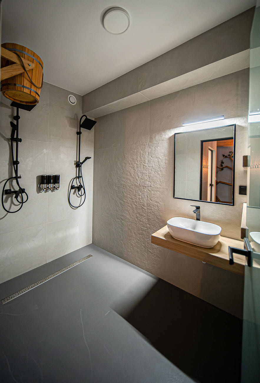 Agroturystyka Za Piecem – Nowoczesna łazienka z kabiną prysznicową typu walk-in, drewnianą deszczownicą kubełkową, przyborami toaletowymi na ścianie i umywalką z lustrem na pływającej drewnianej toaletce. Idealna do agroturystyki dla rodziny, charakteryzująca się minimalistycznym designem w neutralnych odcieniach i teksturowanymi ścianami.