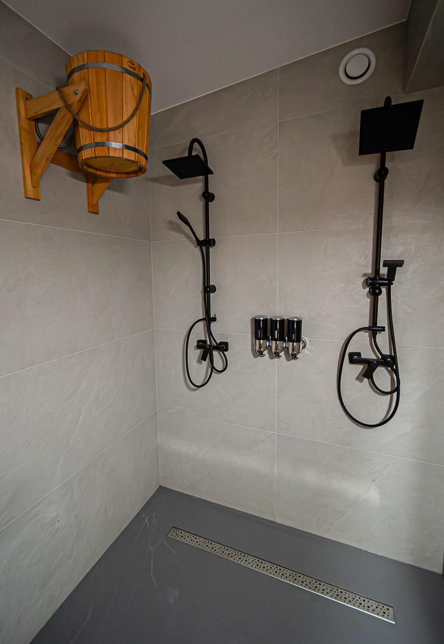 Agroturystyka Za Piecem – Nowoczesny prysznic z dwiema czarnymi głowicami, naściennymi dozownikami mydła i drewnianym wiaderkiem na lewej górnej ścianie, zestawiony z jasnoszarymi kafelkami na ścianach - idealny dla agroturystyki dla rodzin poszukujących komfortu i stylu.