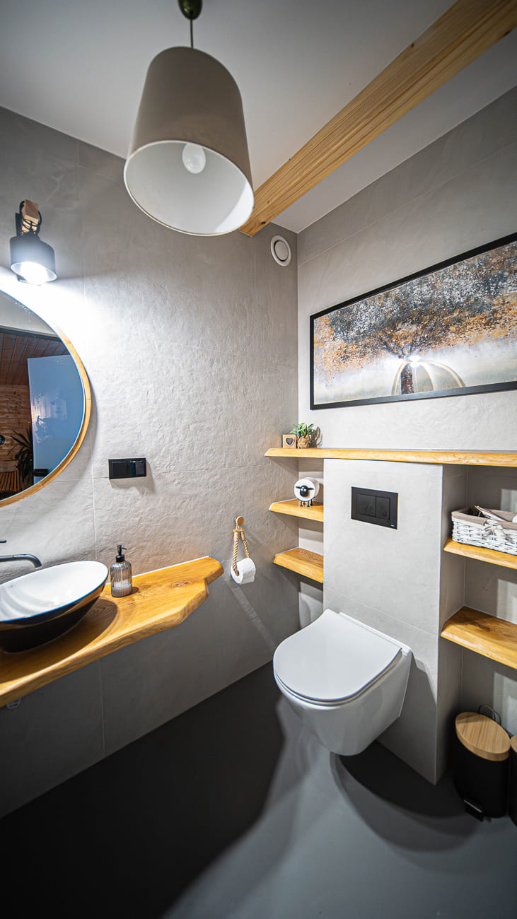 Agroturystyka Za Piecem – Nowoczesna łazienka z pływającą toaletą, okrągłym lustrem nad umywalką, drewnianymi półkami, oświetleniem ściennym, lampą wiszącą i oprawioną grafiką na jasnoszarej ścianie - idealna do spa na wsi lub noclegi dla rodzin z dziećmi.