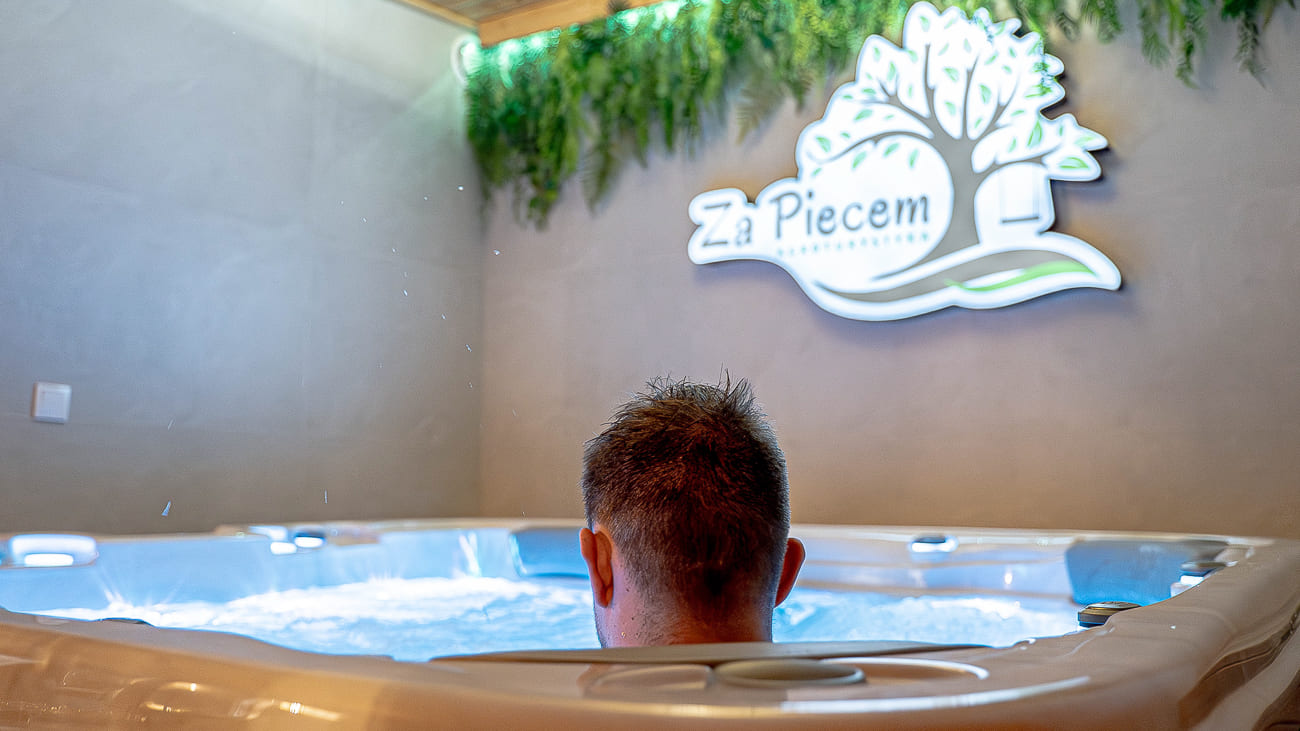 Agroturystyka Za Piecem – Osoba z krótkimi włosami relaksuje się w bąbelkowej wannie z hydromasażem. Zielone rośliny wiszą na ścianie nad świecącym napisem Za Piecem, tworząc spokojną, przypominającą spa atmosferę idealną na wypoczynek na wsi.
