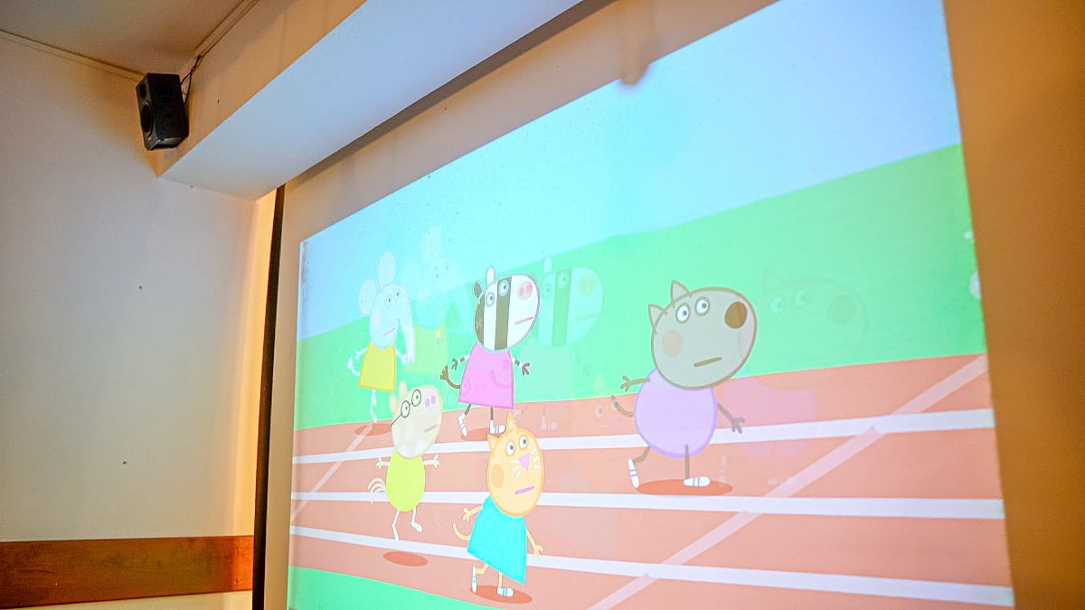 Agroturystyka Za Piecem – Kreskówkowy obraz zwierzęcych postaci biegnących po torze jest wyświetlany na białej ścianie w pomieszczeniu, idealny do agroturystyki dla rodziny. Postacie ubrane są w kolorowe stroje na tle zielonej trawy i błękitnego nieba.