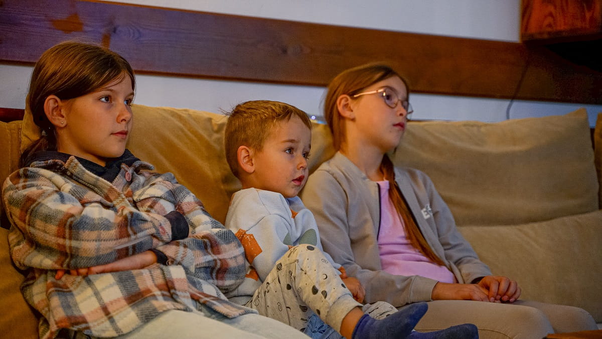 Agroturystyka Za Piecem – Trójka dzieci siedzi blisko siebie na beżowej kanapie w przytulnej agroturystyce dla rodziny, oglądając coś poza ekranem. Wyglądają na spokojne i skupione, z neutralnymi wyrazami twarzy. Tło stanowi drewniana ściana i delikatne oświetlenie.