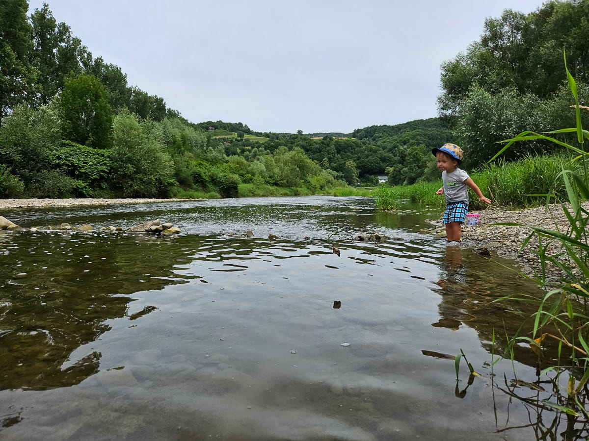 Agroturystyka Za Piecem – Małe dziecko w niebieskim kapeluszu stoi na brzegu płytkiej rzeki, otoczone zielenią i drzewami w pochmurny dzień - idealny moment na agroturystykę dla rodzin poszukujących spokojnej przyrody i łagodnych wód.