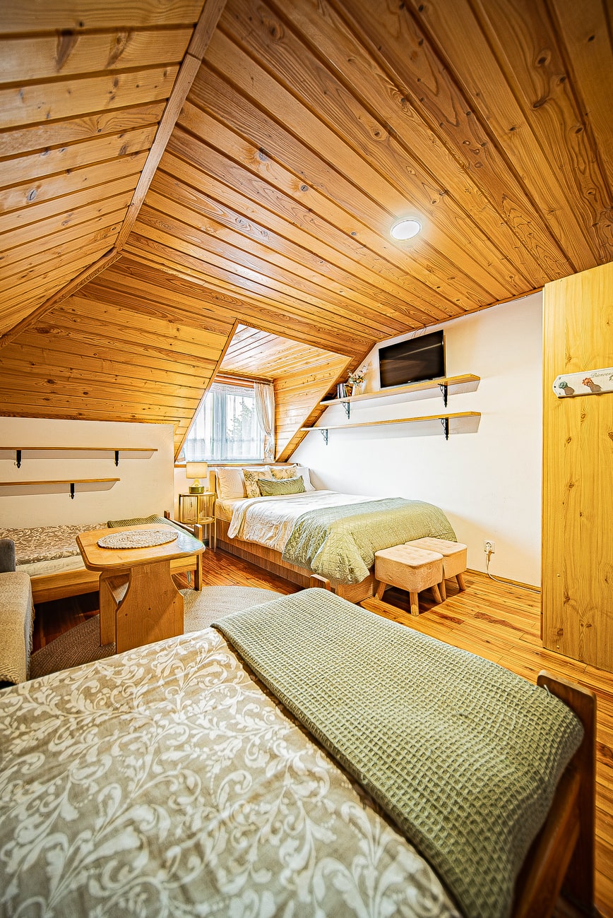 Agroturystyka Za Piecem – Przytulna sypialnia na poddaszu w agroturystyce z dziećmi, z trzema pojedynczymi łóżkami, drewnianymi ścianami i sufitem, małym stolikiem z krzesłami, oknem z zasłonami, półkami, wzorzystą pościelą, zielonymi narzutami i zamontowanym telewizorem.